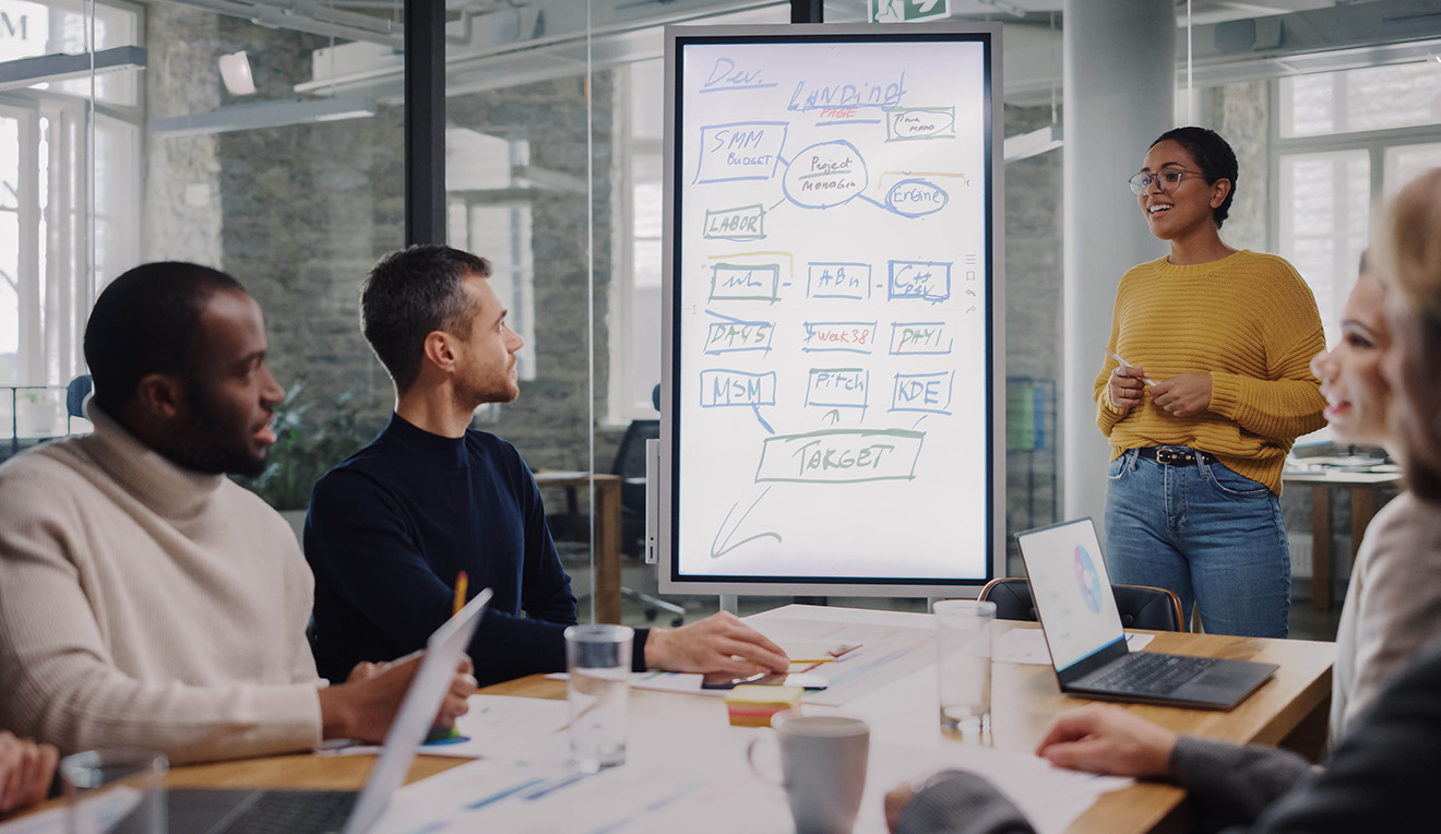 People sit around a table and collaborate while one person stands at a white board drawing a flow chart.