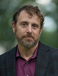 man in a suitcoat and collared shirt looking directly at the camera. Greenery behind him