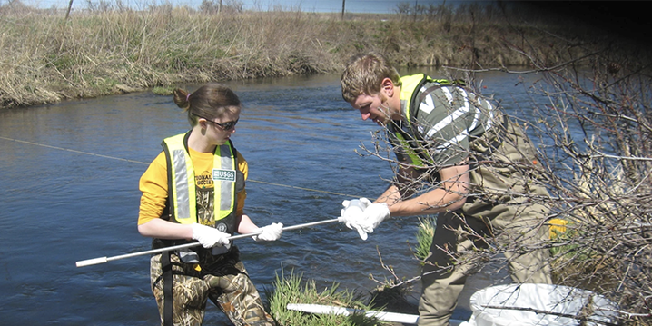Two researchers in wading pants and life vests stand next to river and hold thin metal pole used for taking water samples.