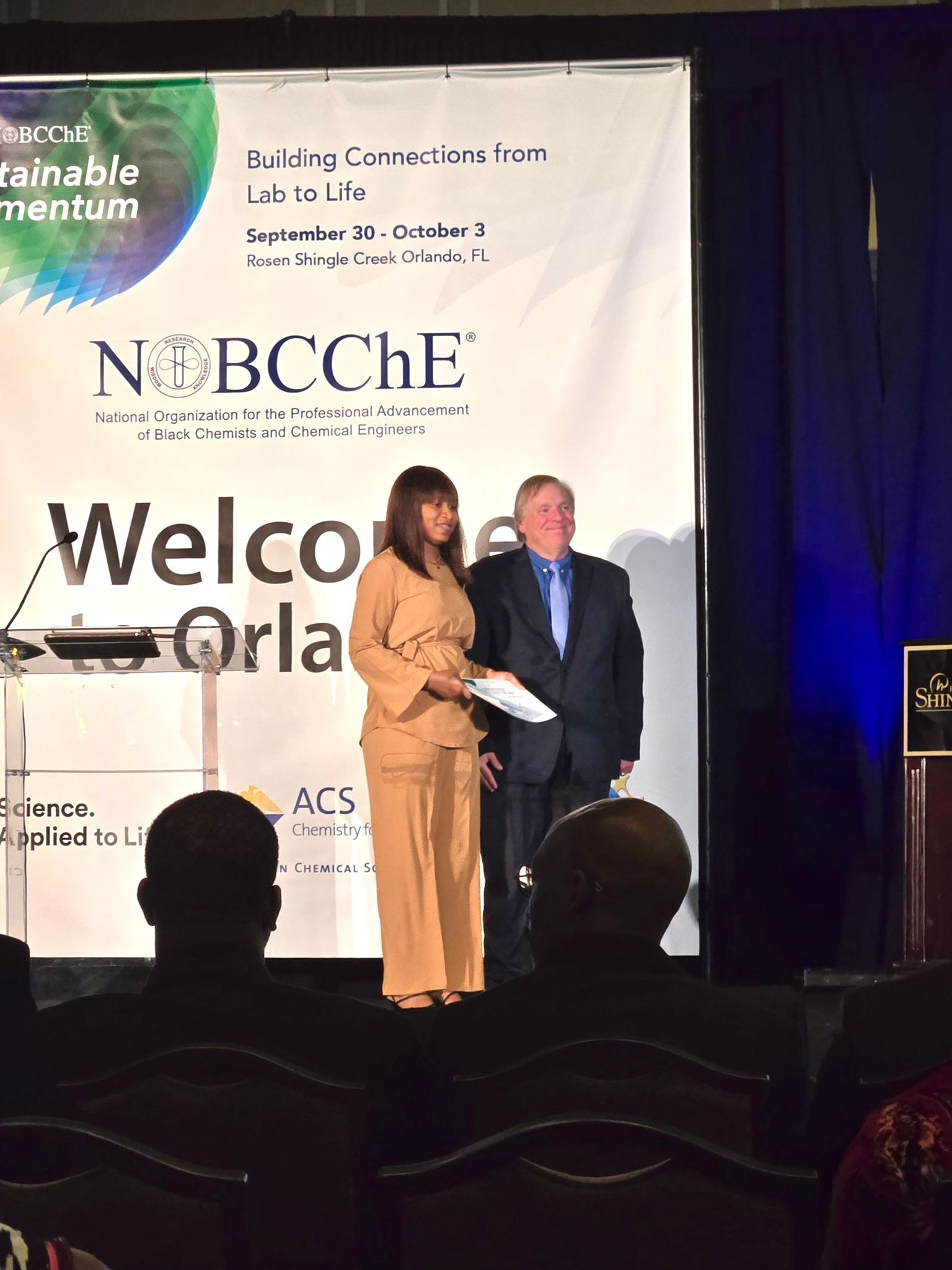 Far shot of Boluwatife Dosunmu accepting Pfizer's award for best graduate research on stage at the NOBCChE conference in front of a backdrop with conference information and branding.