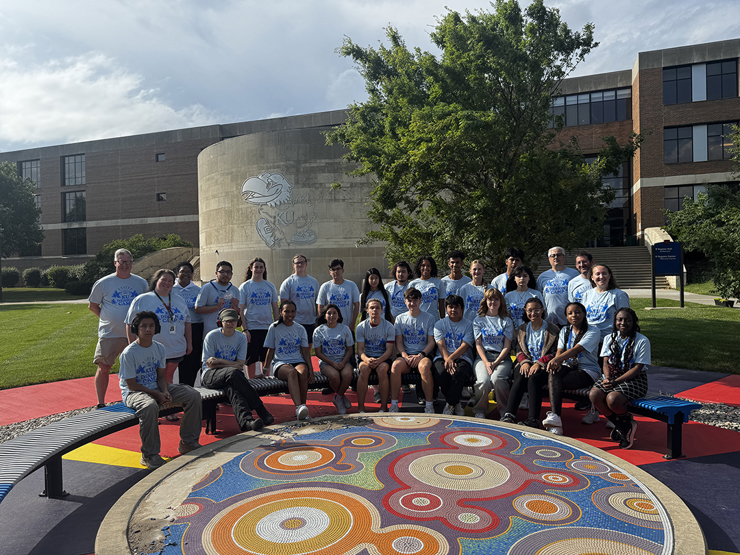Group of people in matching blue T-shirts posing outdoors at the University of Kansas, standing around a colorful circular art installation.
