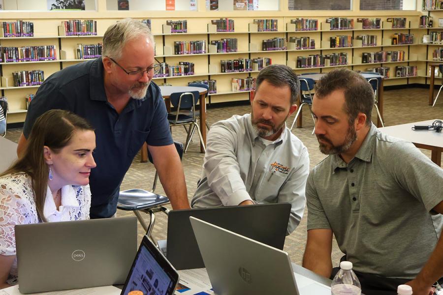 Four educators collaborate around laptops in a library during a professional development session. One person is standing and pointing at a screen while the others focus attentively, highlighting teamwork and engagement in a learning environment.