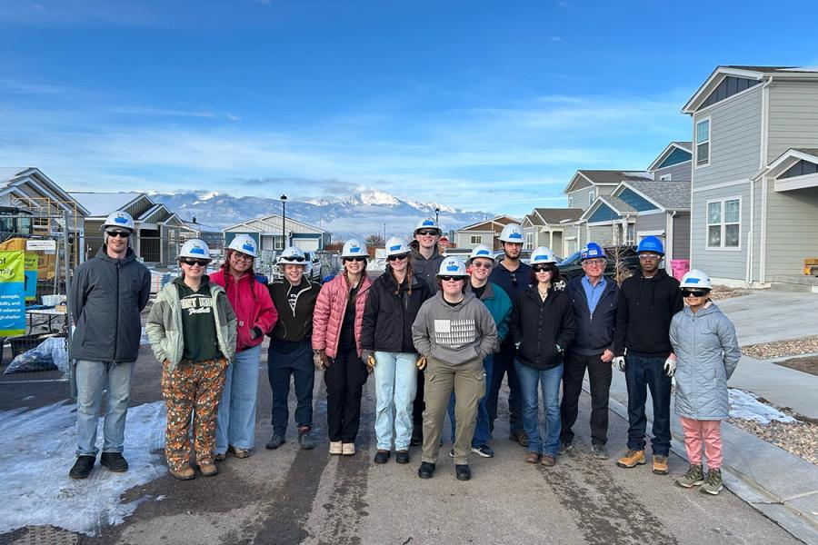 Group of smiling students wearing hard hats and winter jackets, standing with homes and mountains in the background.
