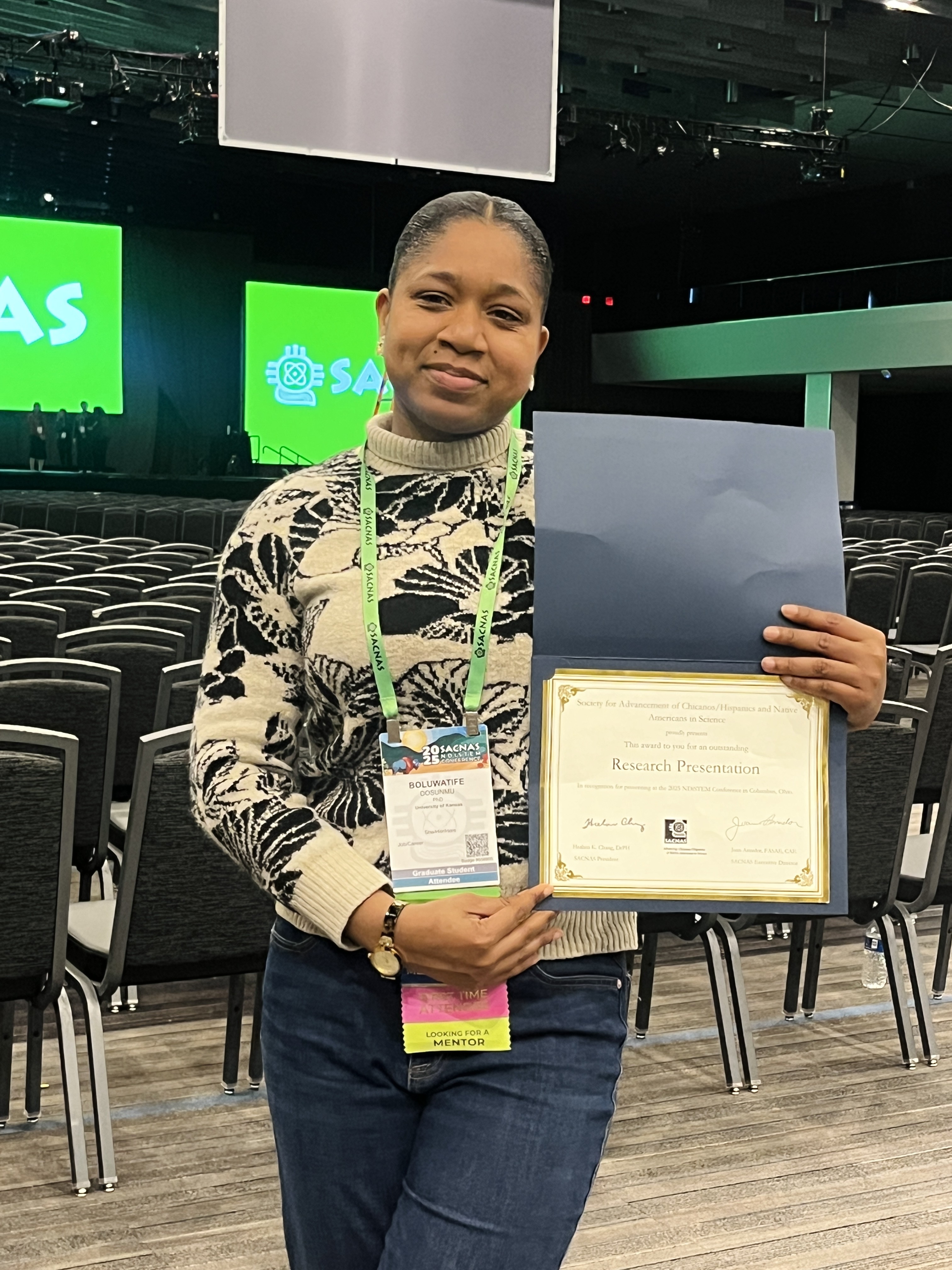 Boluwatife holds her Research Presentation award, with the SACNAS conference stage visible in the background