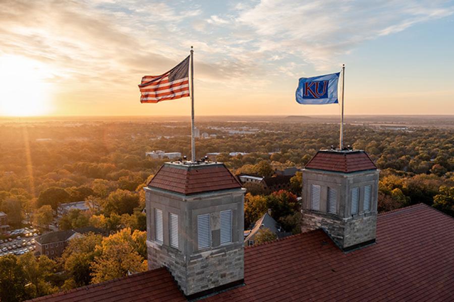 The American flag and KU flag atop Fraser Hall wave in the wind as the sun rises in the background.