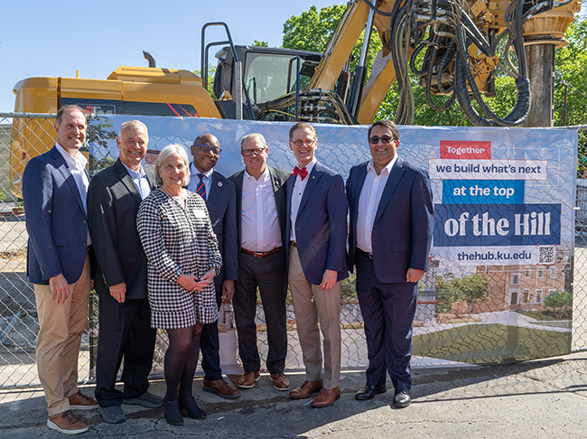 Brian Anderson, John and Cindy Hill, Dean Jide Wintoki, Chancellor Douglas Girod, Endowment President Dan Martin and Provost Arash Mafi stand in front of machinery and a banner reading "Together we build what's next at the top of the Hill, thehub.ku.edu" at the construction site