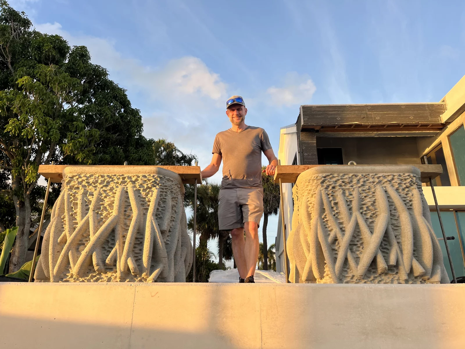 Keith Van de Riet stands on a waterfront walkway between two mangrove inspired concrete seawall planters, featuring textured surfaces that mimic mangrove roots, with trees and buildings in the background at sunset.