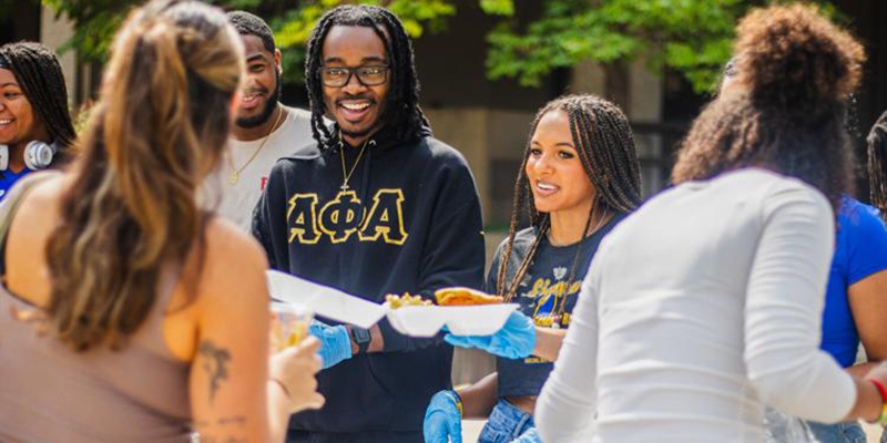 A group of people are gathered outdoors at a food serving event where one individual is wearing a black hoodie with gold Greek letters. Two people are serving food, wearing gloves, and another person is holding a plate.