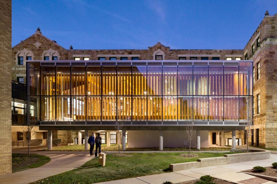Exterior view of John C. Gaunt Fourm at dusk, featuring vertical wood slats inside, illuminated interior lighting, and a landscaped courtyard with a walkway and two people walking below the structure.