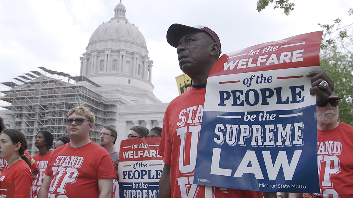 A photograph of a man holding a sign outside of the Missouri Capital. He is wearing a red T-shirt and holding signs made by Stand Up KC, an organization advocating for better pay for restaurant workers.