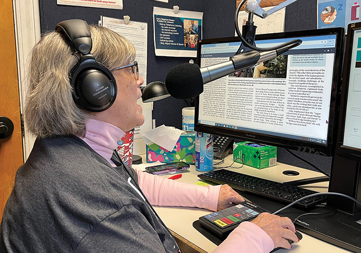 Dee Dee Eccles has short grey hair, wears glasses, and has headphones on as she speaks into a microphone at the Audio-Reader studios with The Leaven on the screen of the computer in front of her.