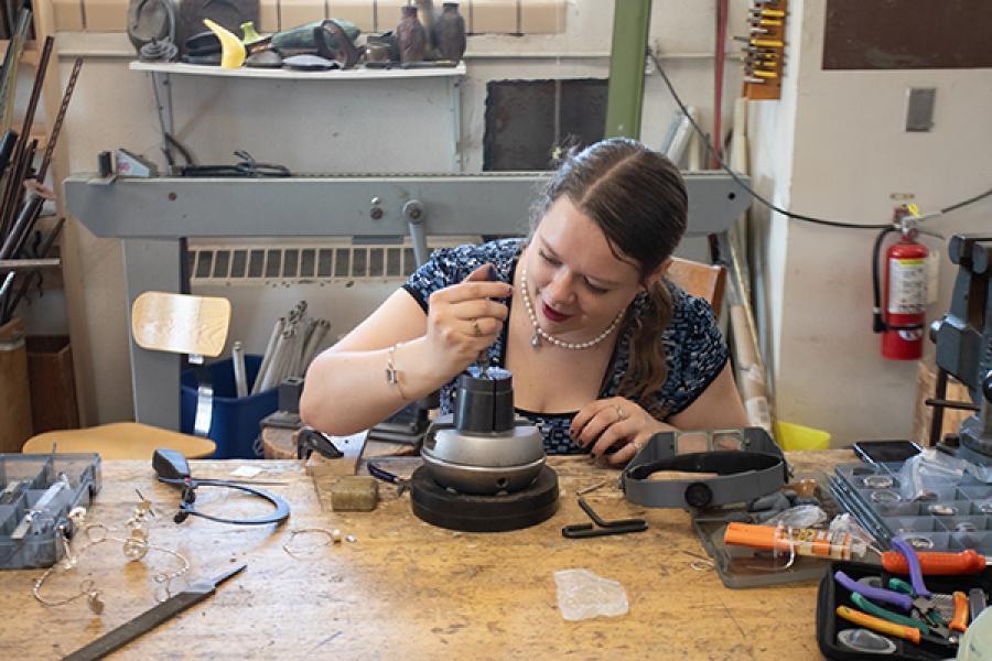 A person is focused on working with jewelry at a craftsman's bench. The table is scattered with tools, wires, and equipment. There's a vise on the right and a wall with various tools in the background.