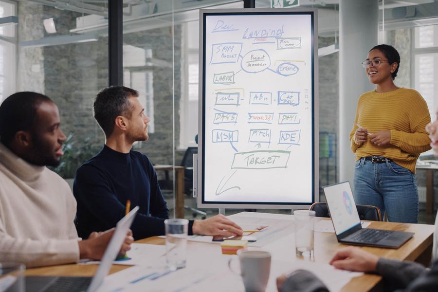 People sit around a table and collaborate while one person stands at a white board drawing a flow chart.