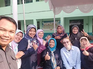 A group of adults smiling and posing for a selfie, some giving a thumbs up, in front of a building.