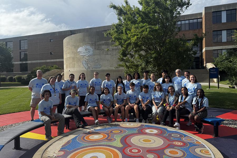 Group of people in matching blue T-shirts posing outdoors at the University of Kansas, standing around a colorful circular art installation.