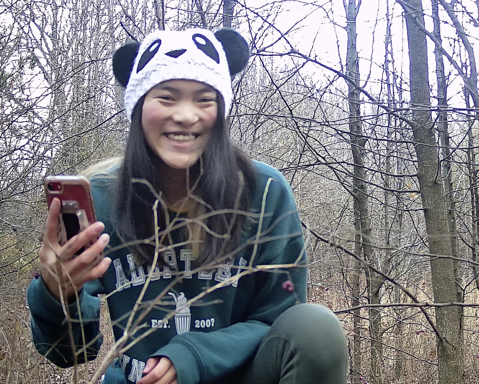 Mira Rausch in wooded area crouching, holding an iPhone and facing the camera; her face is somewhat blurred because the camera is triggered by motion.