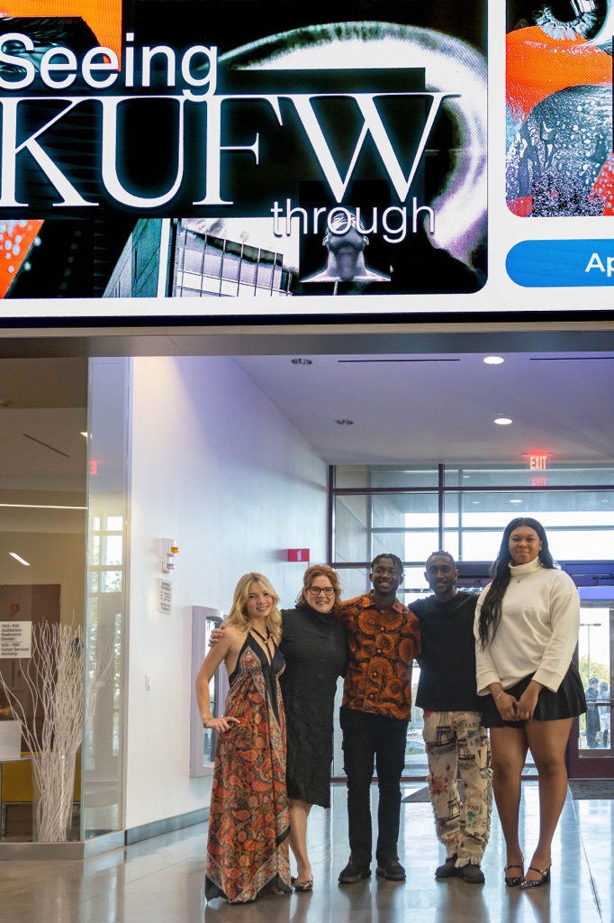 A group of five people pose for a photo under a gaint videoboard advertising KUFW 'Seeing through KUFW,' featuring close-up graphics of an eye and urban scenery in a building lobby.