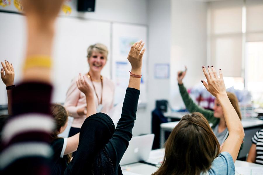 A photo of young students raising their hands in a classroom with a teacher standing near the front of the room.