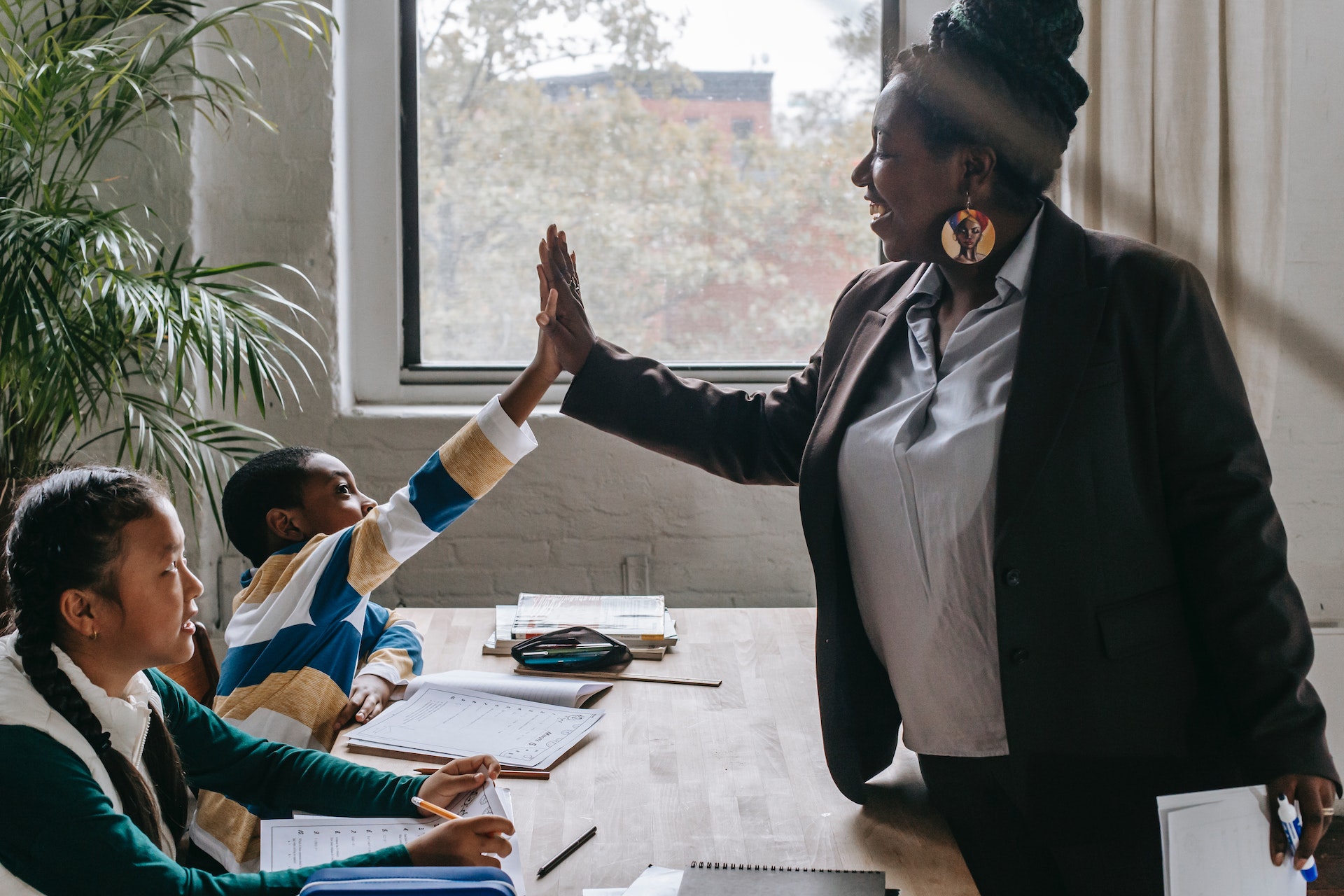 "A woman teaching two students at their desk gives a high five to one boy sitting next to a girl"