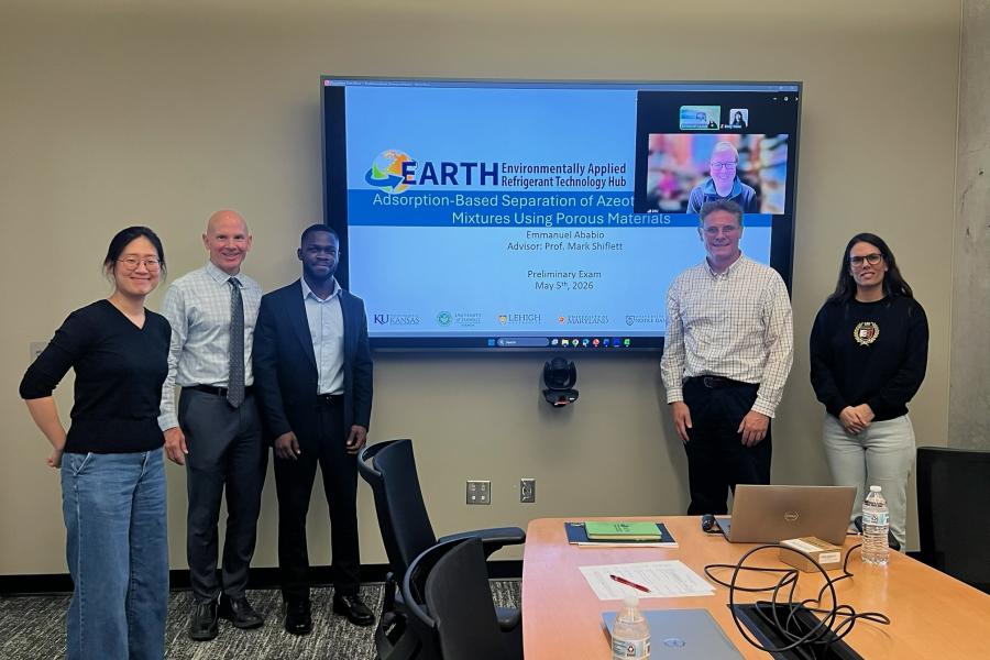 Five people stand in front of a projector screen showing a preliminary exam presentation for a PhD student and a small Zoom window with an additional professor attending virtually