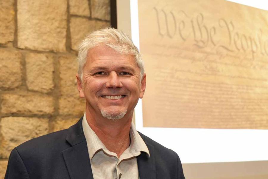 KU researcher Chris Koliba standing in front of background with "We the People ..."