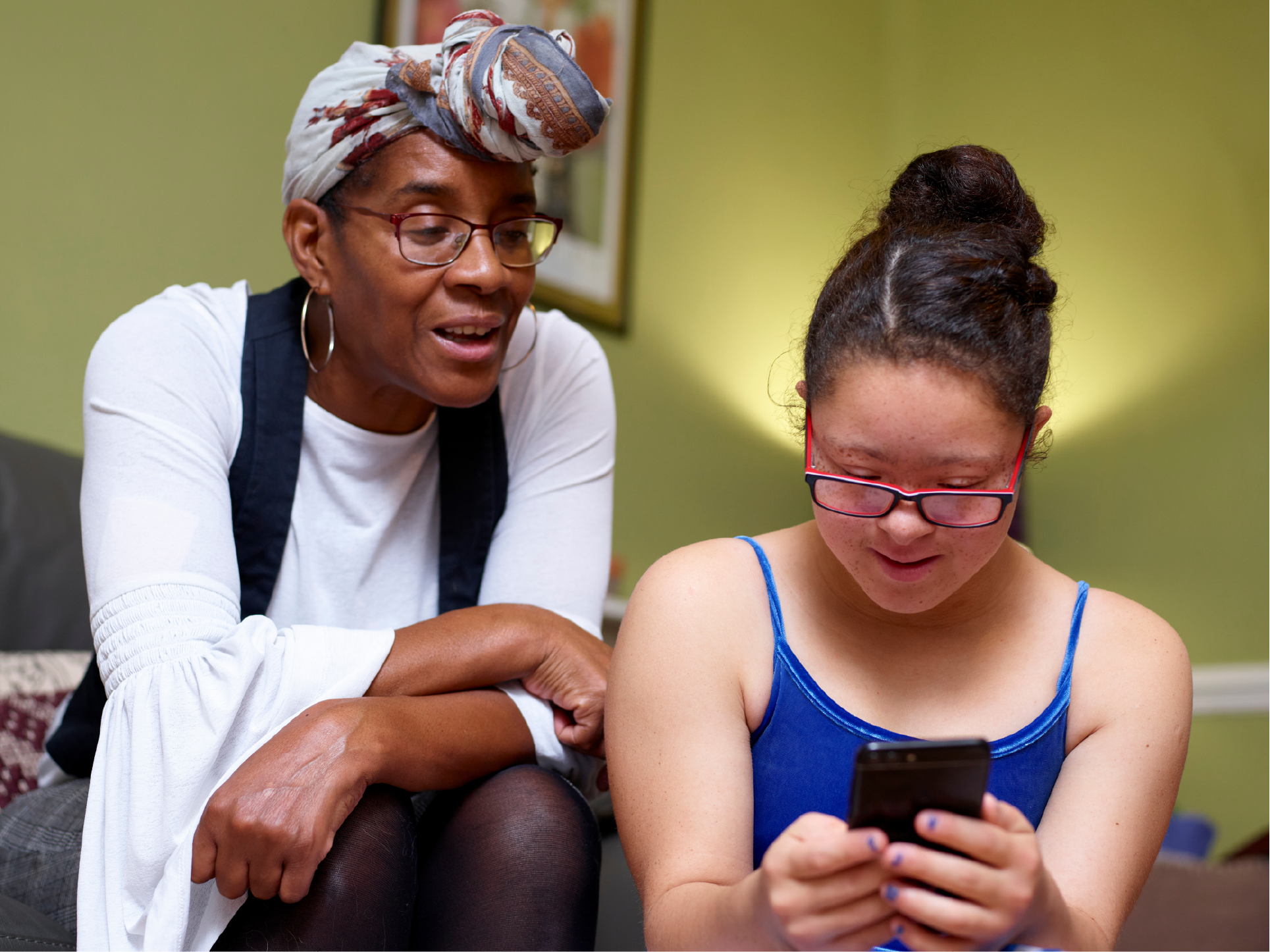 "An older woman with her hair tied in scarf speaks to a younger woman holding a phone"