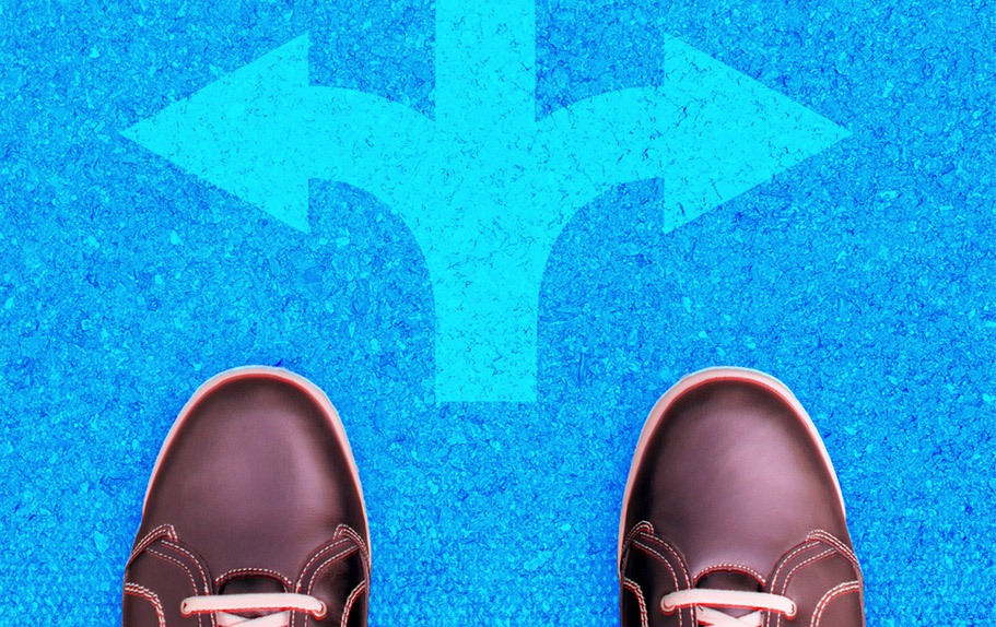 A pair of feet stand on pavement at crossroads marked by painted white arrows pointing in different directions