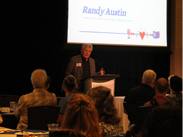 Randy wears a black button up shirt under a patterned suit coat.