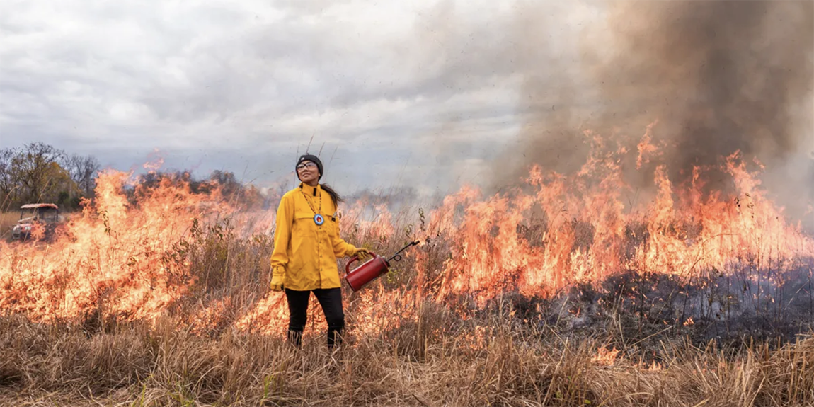 Person in yellow fire jacket in front of controlled prairie burn.