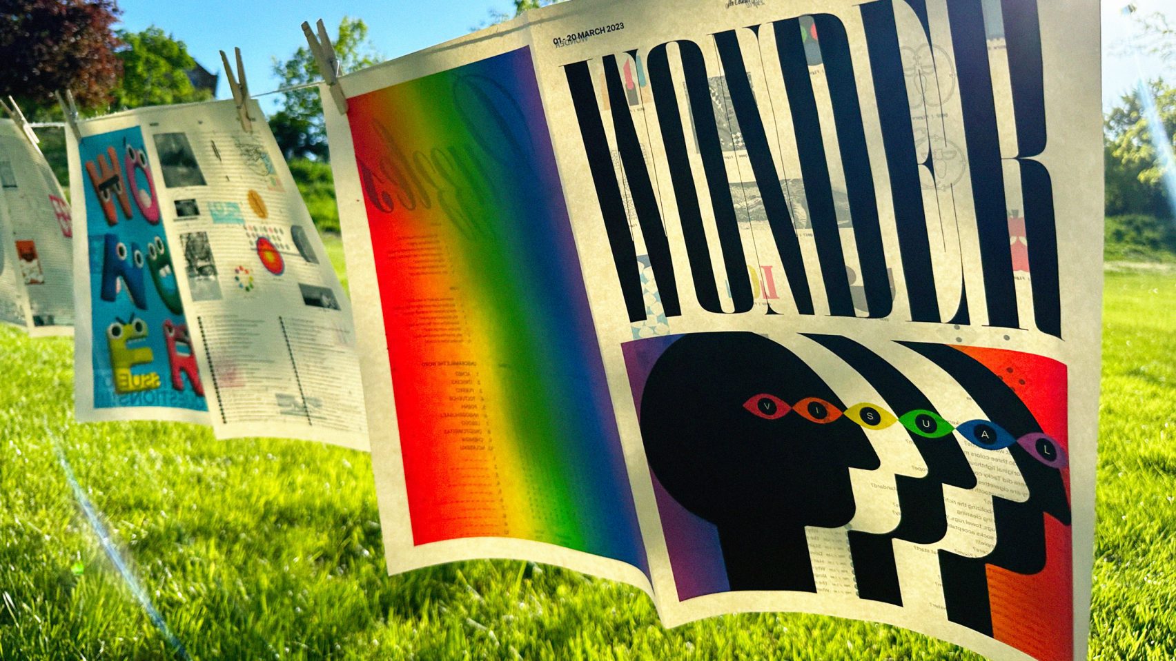 A photograph of sheets of newspaper hanging from pegs on a string line in a green space, with multicolored details on them