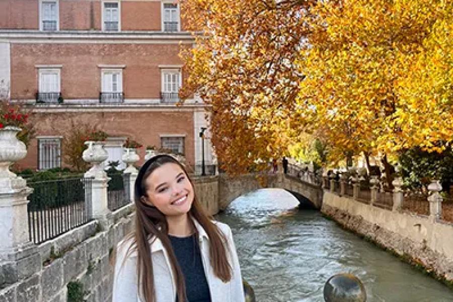 A woman wearing a long white coat stands on a bridge overlooking a canal with autumn foliage in the background.