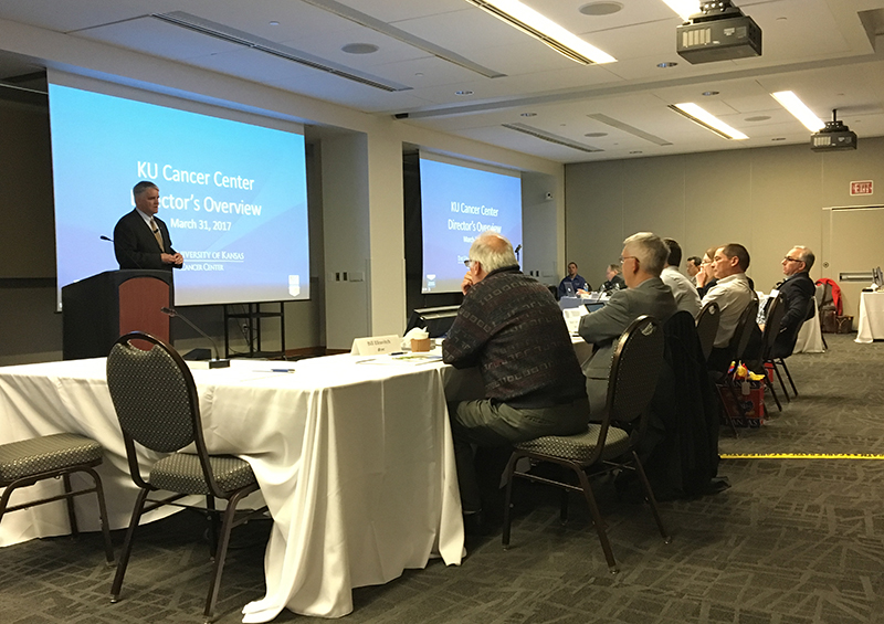 A presenter speaking at the front of the room to multiple rows of tables filled with attentive listeners.