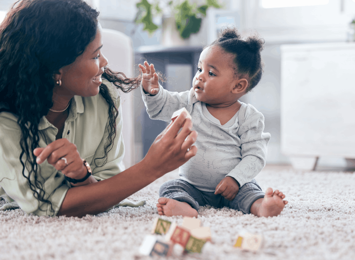 A baby sitting on a carpeted floor reaches toward her mother, who is smiling at her and holding a toy block.