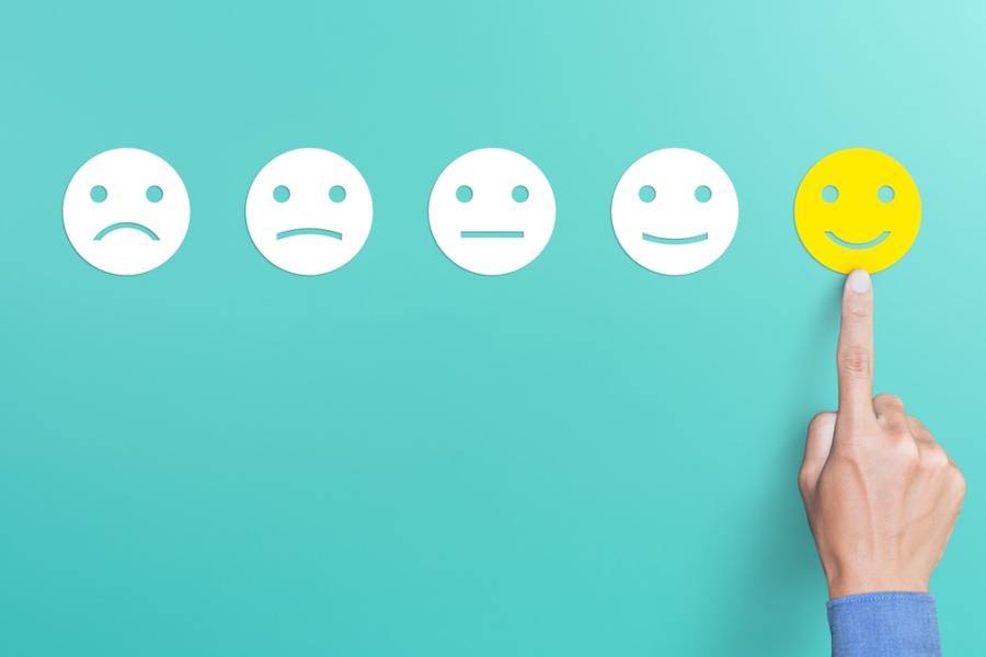 Five smiley face cutouts ranging from unhappy to happy are lined up, with a human hand pointing to the happiest one.