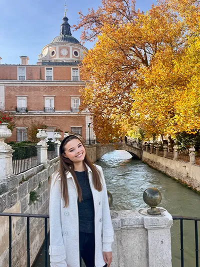 A woman wearing a long white coat stands on a bridge overlooking a canal with autum foilage in the background.
