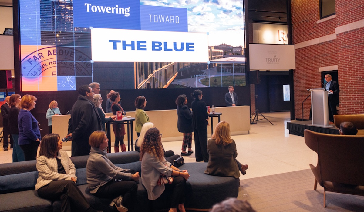 Chancellor Douglas A. Girod speaking to group at Jayhawk Welcome Center with large screen displaying "Towering Toward the Blue."