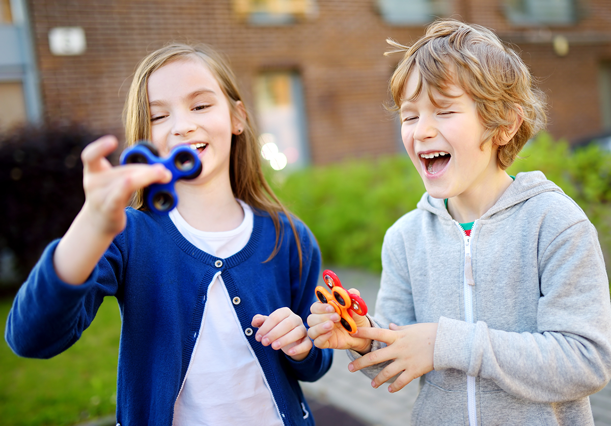 A boy and a girl smile and play with fidget spinners outdoors together.