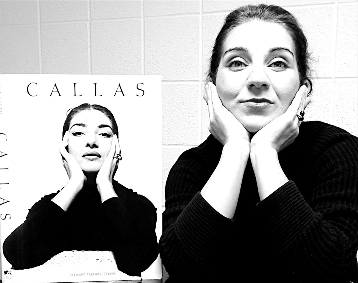 Stella Markou leans forward with chin on hands, emulating pose in photo of Maria Callas on easel next to her.