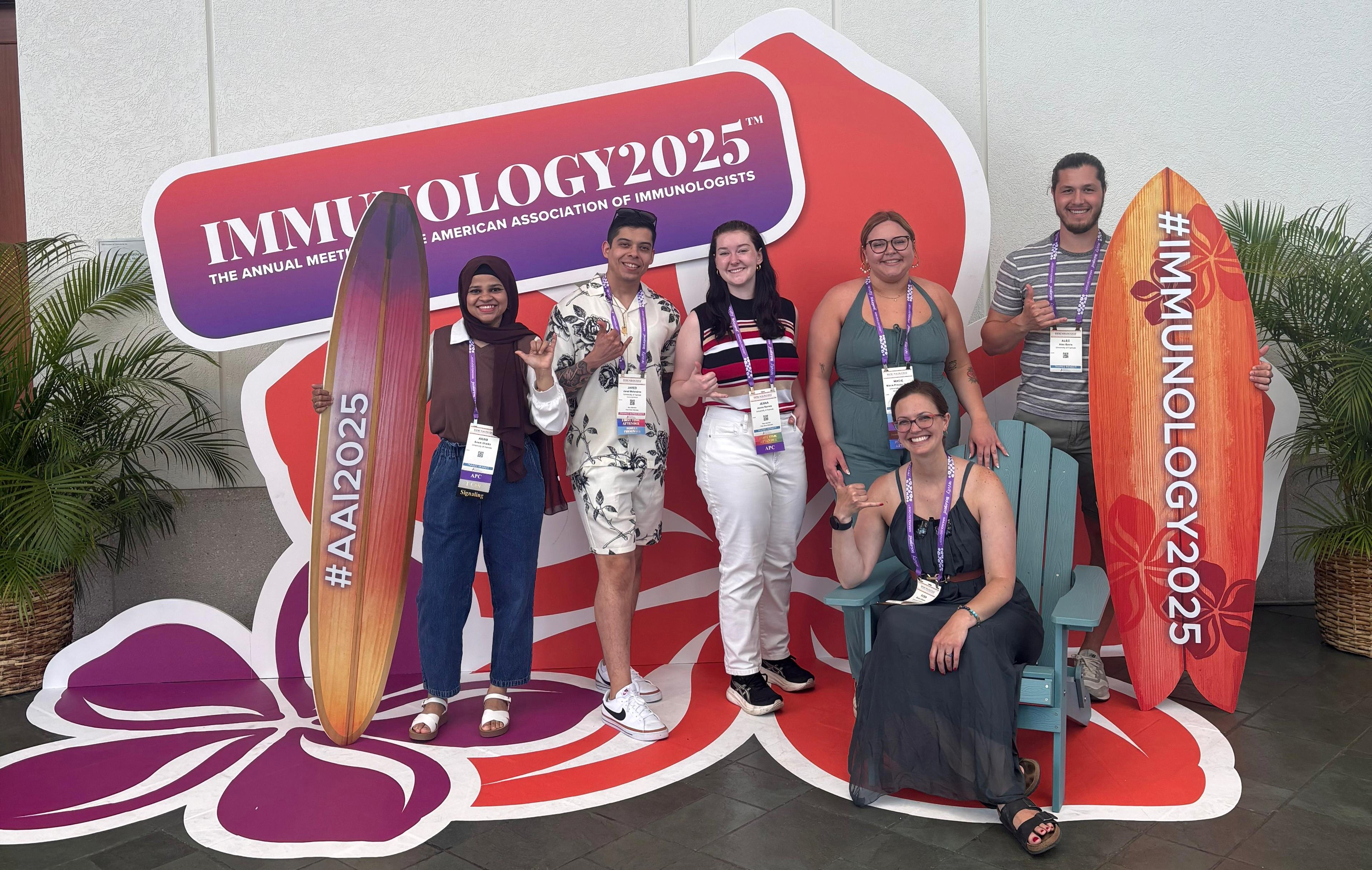 Anam Shaikh, Jared Melendrez, Jenna Barnes, Macie Proctor-Roser, Alec Bevis, wand Dr. Robin Orozco post with surfboards reading #immunology2025 in a Hawaii themed photo backdrop