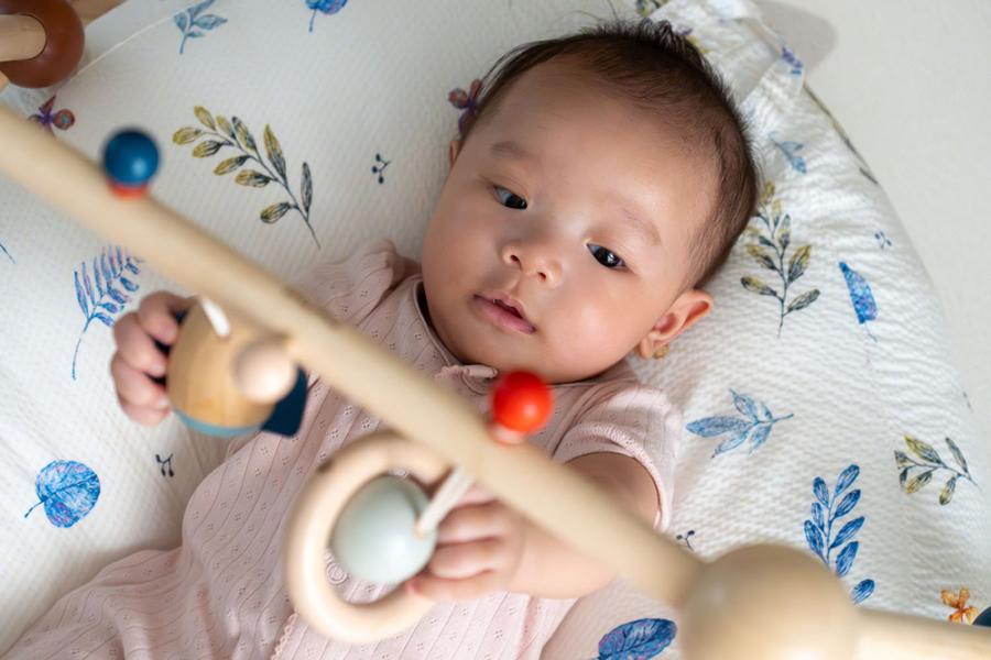 A baby with dark hair wearing a pink outfit reaches for toys hanging from a mobile hanging overhead her crib