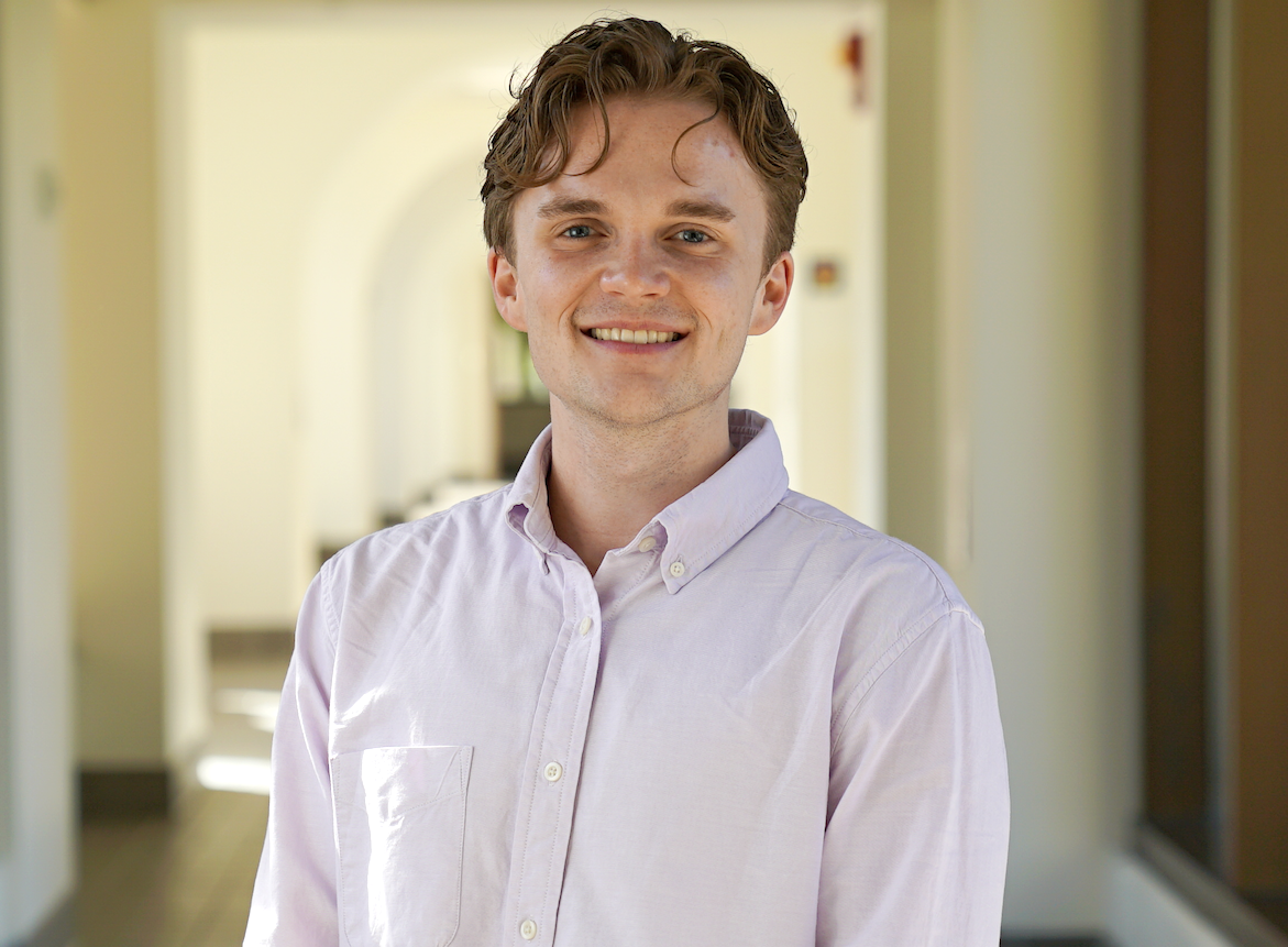 "A man with light brown hair and a white shirt stands in a hallway while smiling