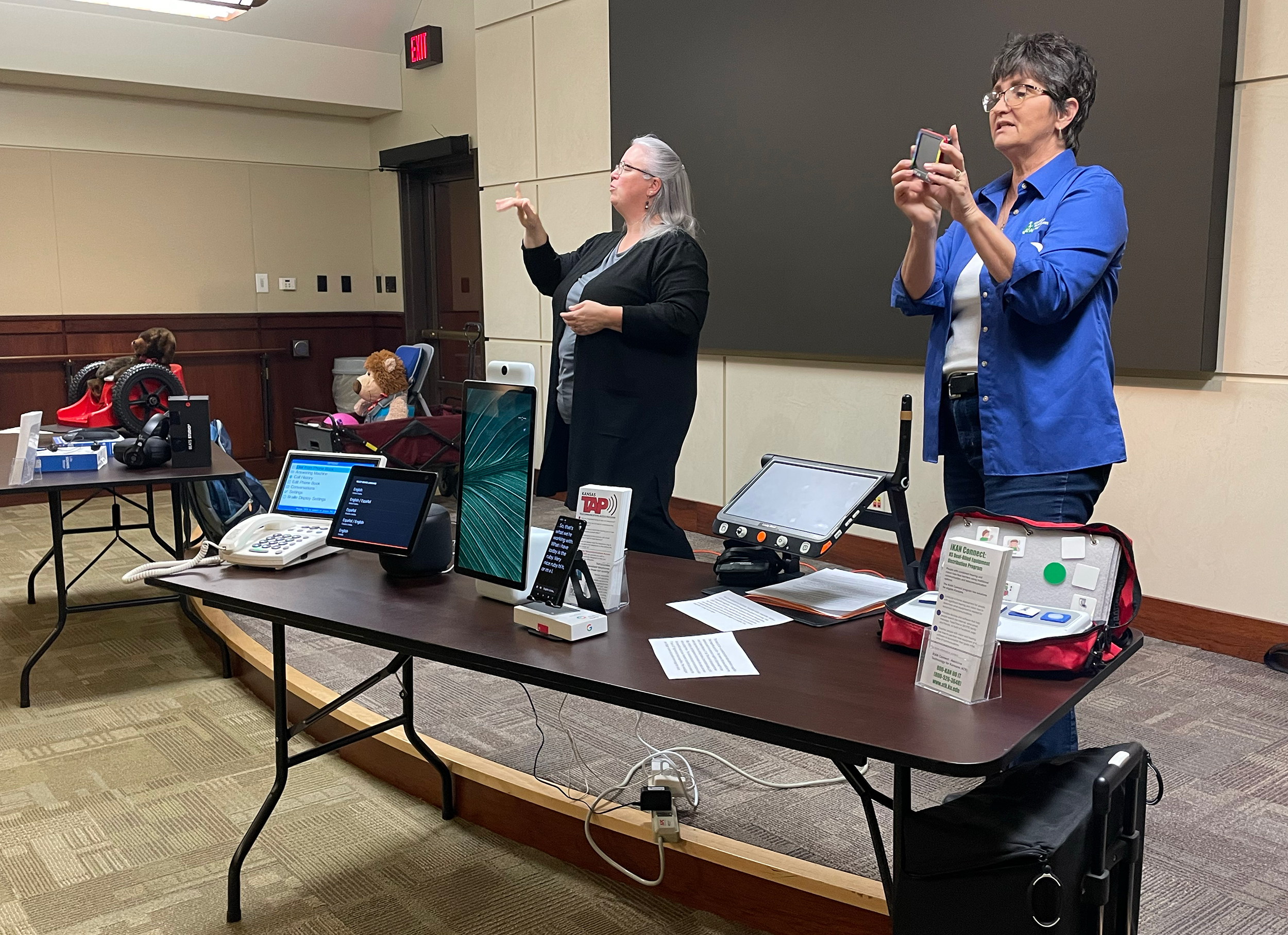 "A woman speaks from a platform with assistive technology on a table in front of her while a woman translate in American Sign Language"