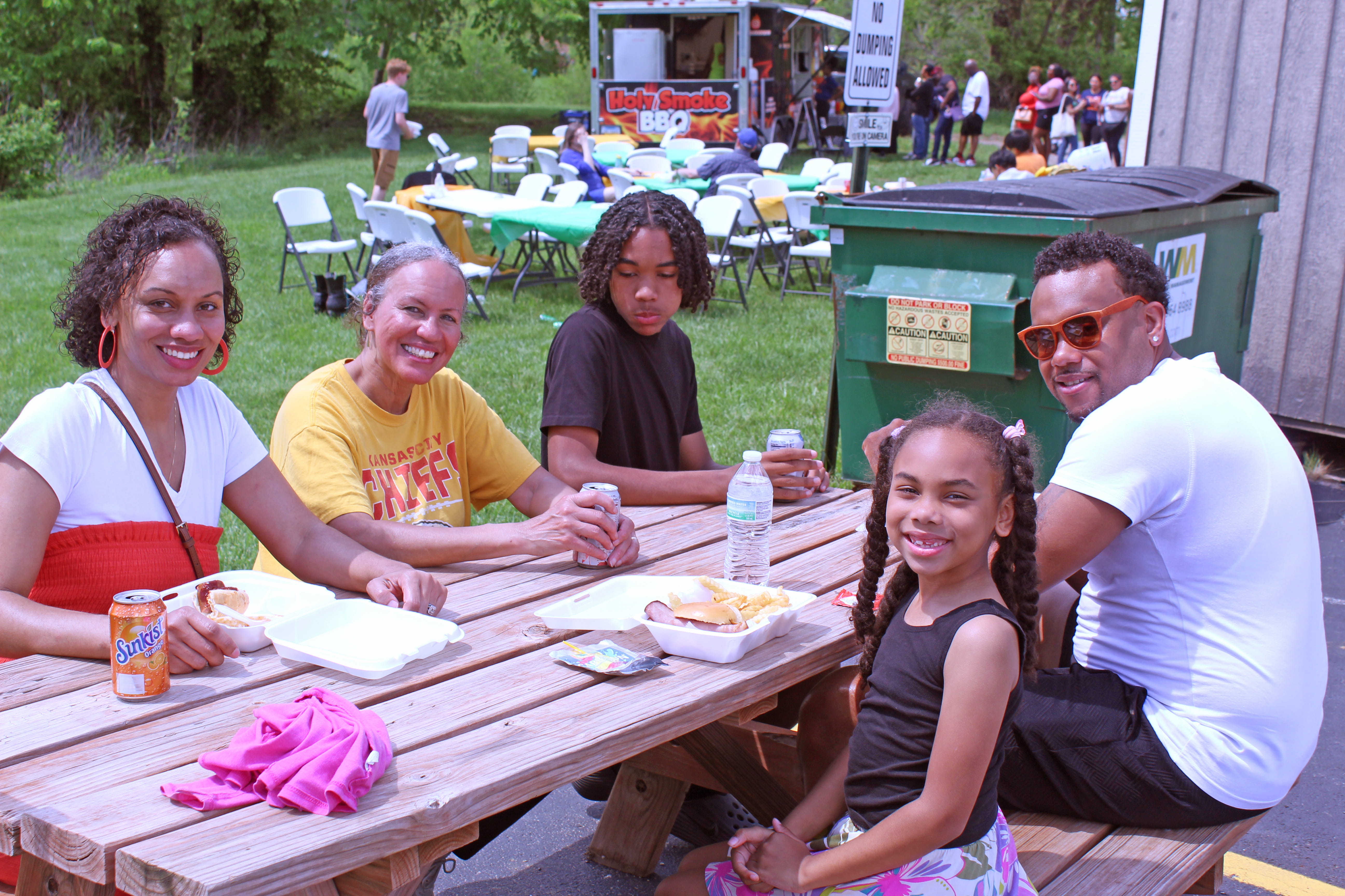 A family of three adults and two children sit at a table with plates of food at a community picnic.