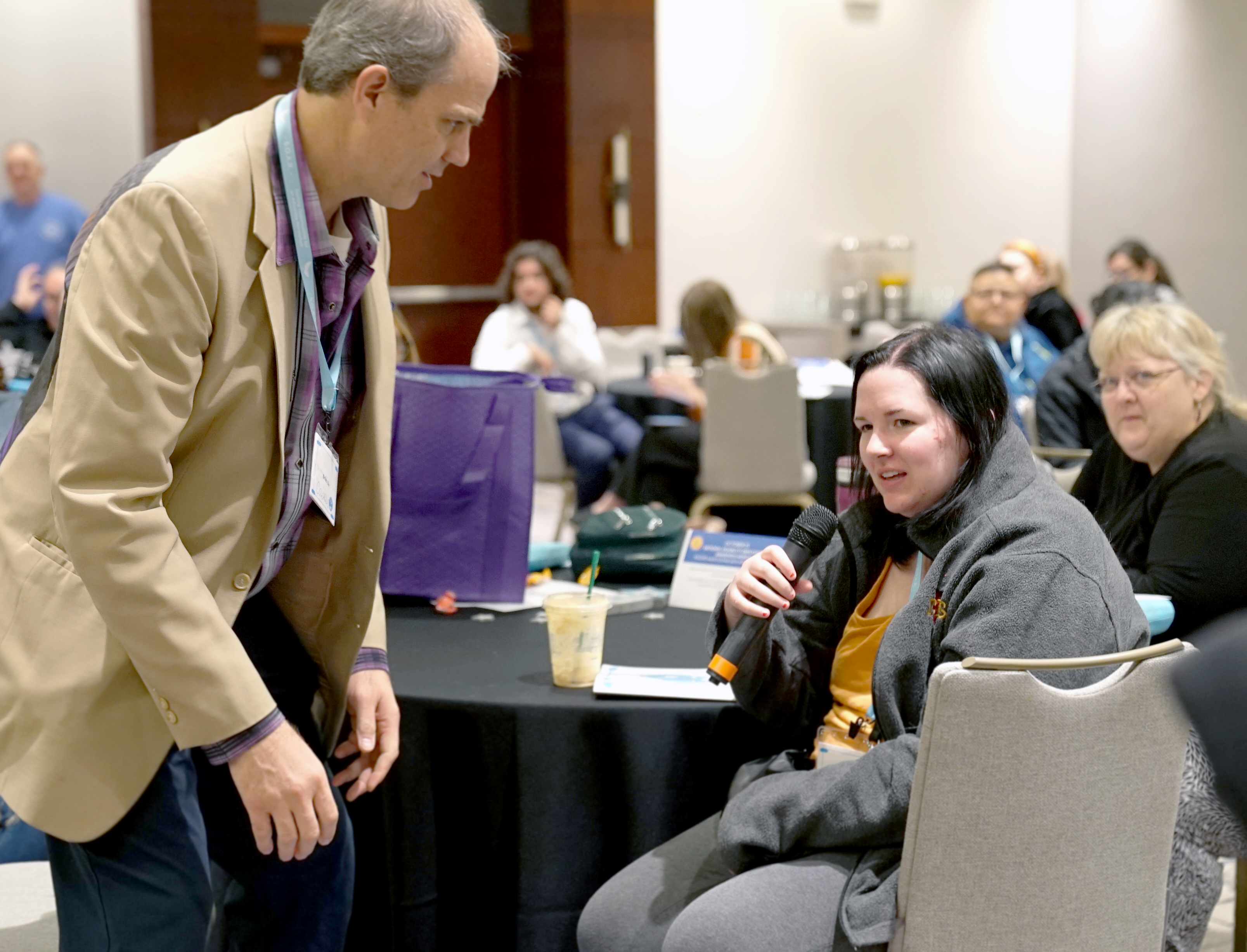 "A woman with straight black hair wearing a gray jacket speaks into a microphone while seated at a table in a conference room"