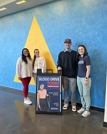 Four students pose in front of a blue with a yellow triangle with an A-frame sign advertising the KU blood drive.