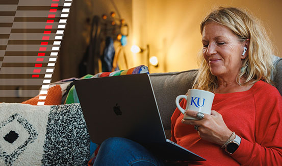 Smiling woman sitting on couch looking at laptop while holding coffee mug with KU logo.