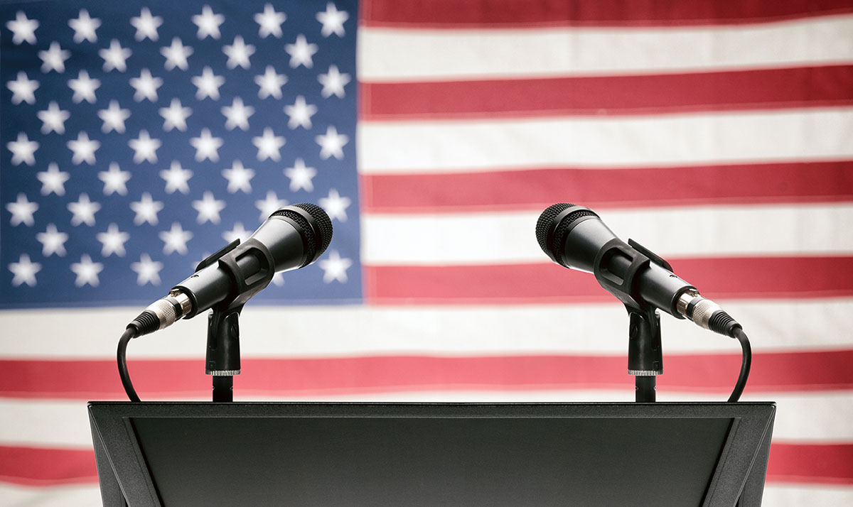 Image of lectern with two microphones with American flag in background