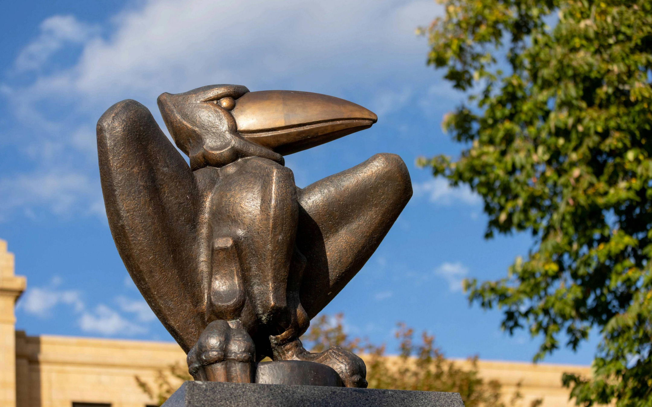 A Jayhawk statue sits in the foreground with a building and blue sky behind it.
