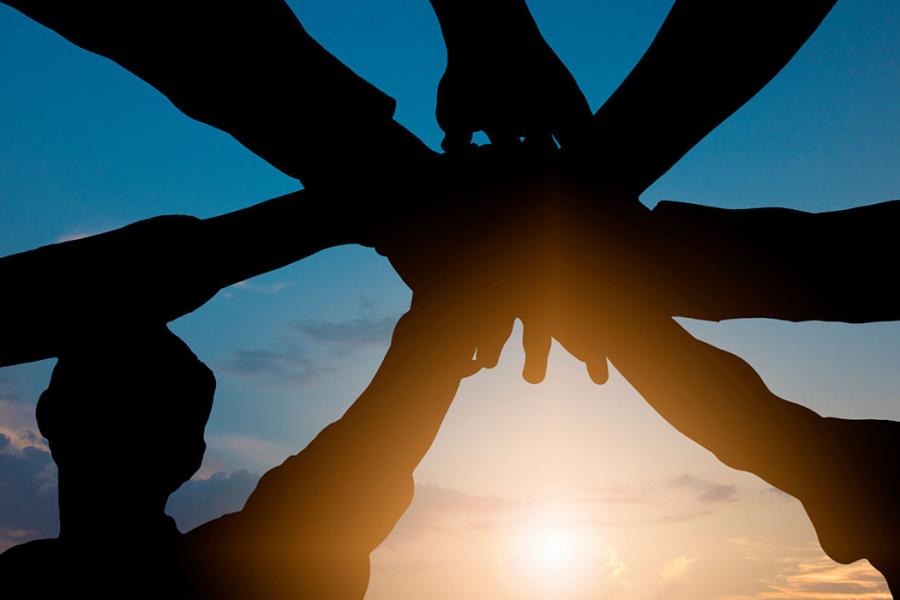 A group of hands are joined before the backdrop of a sunset.