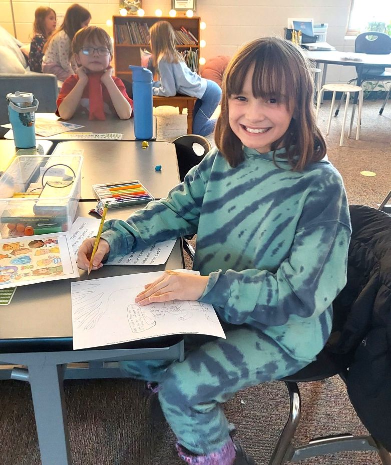 Student smiling and sitting at a group desk with a coloring page.